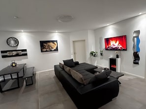 A modern living room with a black sofa, a flat-screen TV, a fireplace, and a dining area with a table and chairs. - Ferienhaus Am Gla Ck (Bad Zwischenahn)