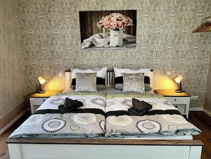 A double bed with a patterned bedspread, two bedside tables with lamps, and a wall-mounted picture of flowers. - Ferienhaus Am Gla Ck (Bad Zwischenahn)