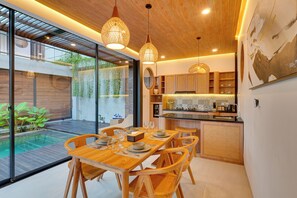 A modern kitchen with a dining area, wooden furniture, and a view of a swimming pool. - Villa Tajung - Umalas (Badung)