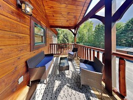 Una terraza de madera con mobiliario de mimbre y una mesa de vidrio, con vistas a un área boscosa.
