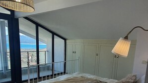 Et soveværelse med en stor seng, et natbord med en bog, en lampe og havudsigt gennem store vinduer.