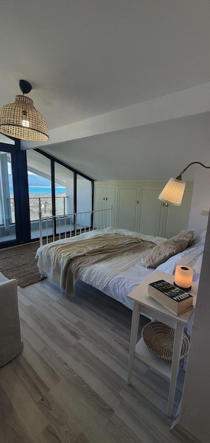 Un dormitorio con una cama grande, una mesita de noche con un libro, una lámpara y vistas al mar a través de amplias ventanas.