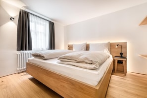 A modern bedroom with a wooden bed, a nightstand with a lamp, and a window with curtains. - Mã¼nsterhaus (Schruns)