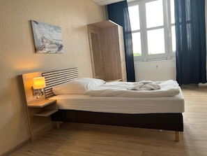 Quarto de hotel com uma cama, uma mesa de cabeceira com abajur, um quadro na parede e uma janela com cortinas.