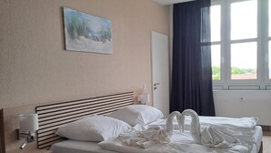 Ein Hotelzimmer mit Bett, Nachttisch, einem Gemälde, einem Fenster mit Vorhängen und Blick auf Grünfläche.