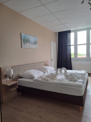 Quarto de hotel com cama, mesa de cabeceira, pintura, janela com cortinas e vista para o verde lá fora.