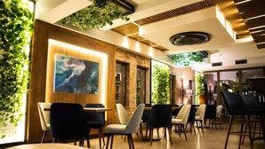 A modern interior with a dining area featuring round tables and chairs, a large abstract painting, and green plants on the walls. - Hotel City In (Kocani)