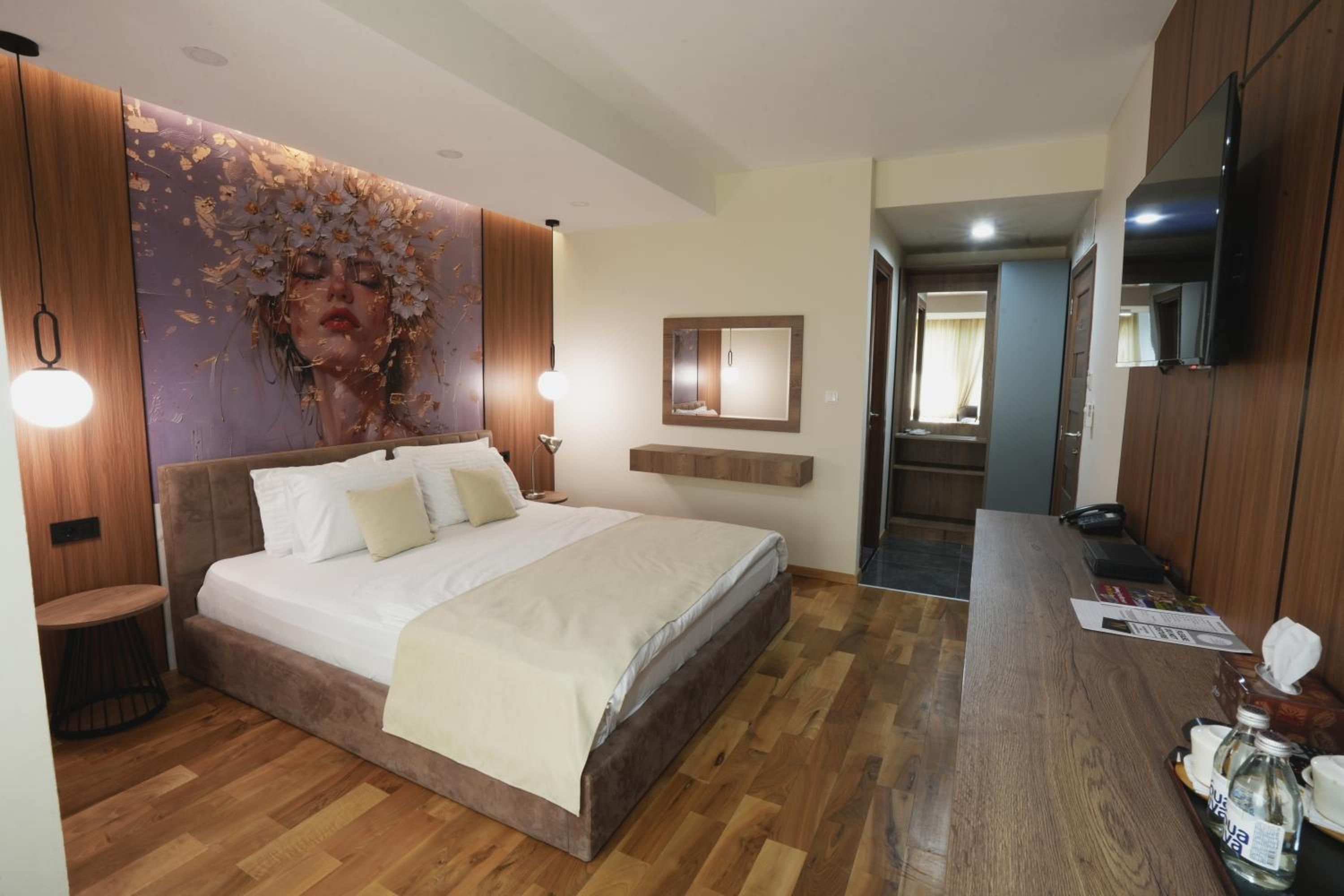 A modern hotel room with a large bed, wooden flooring, a mounted TV, and a bathroom visible through an open door.