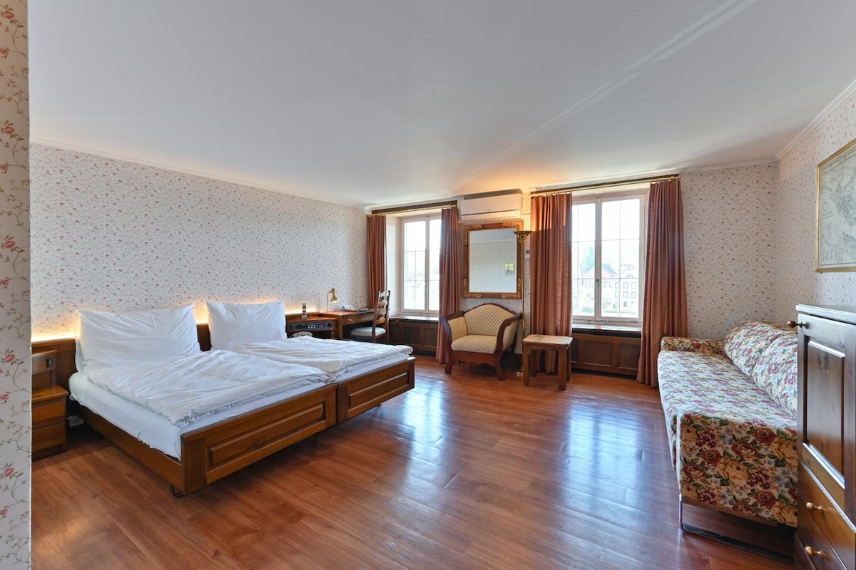 Habitación de hotel con cama doble, un escritorio pequeño con silla, un sofá y un ventanal con cortinas.