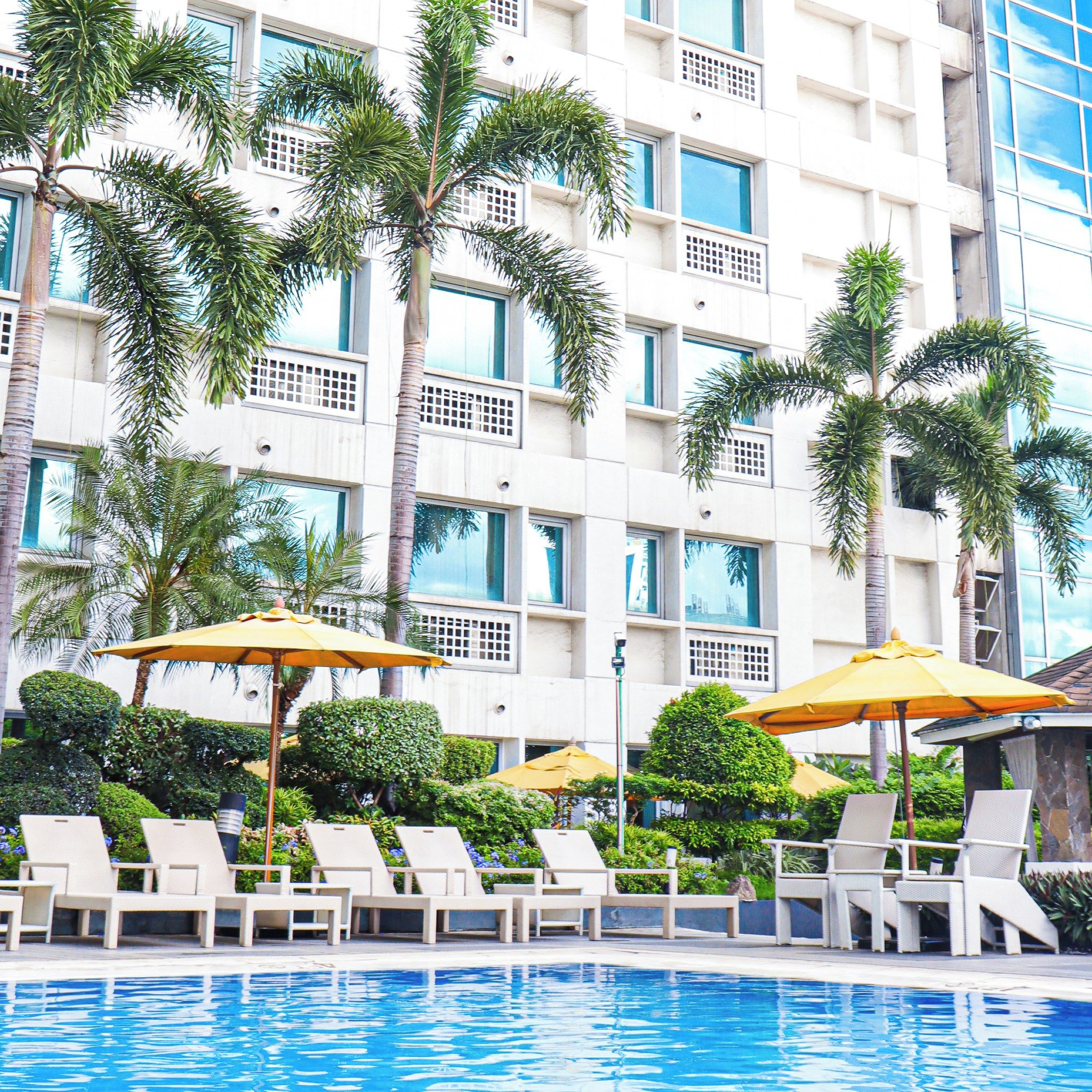 A hotel pool area with lounge chairs and umbrellas, surrounded by palm trees and a building with multiple windows.