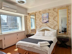 A hotel room with a bed, a nightstand, a mirror, and a window with a view of buildings. - Grand Cenia Residence with 2 Bedrooms Condo Unit (Cebu City)