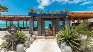 Una entrada frente a la playa con un paseo techado que conduce a una zona de estar, carteles para una escuela y un club de playa, y vistas al océano.