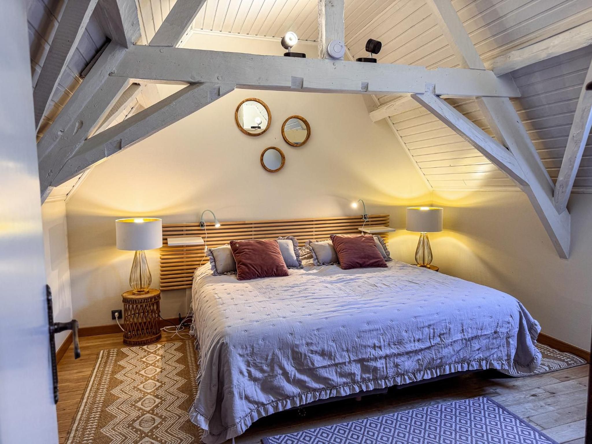 Een slaapkamer met schuine balkenplafond, houten balken, een bed met hoofdeinde, twee bedlampen en een kamerbreed tapijt met dessin.