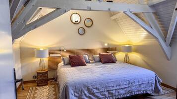 Een slaapkamer met schuine balkenplafond, houten balken, een bed met hoofdeinde, twee bedlampen en een kamerbreed tapijt met dessin.