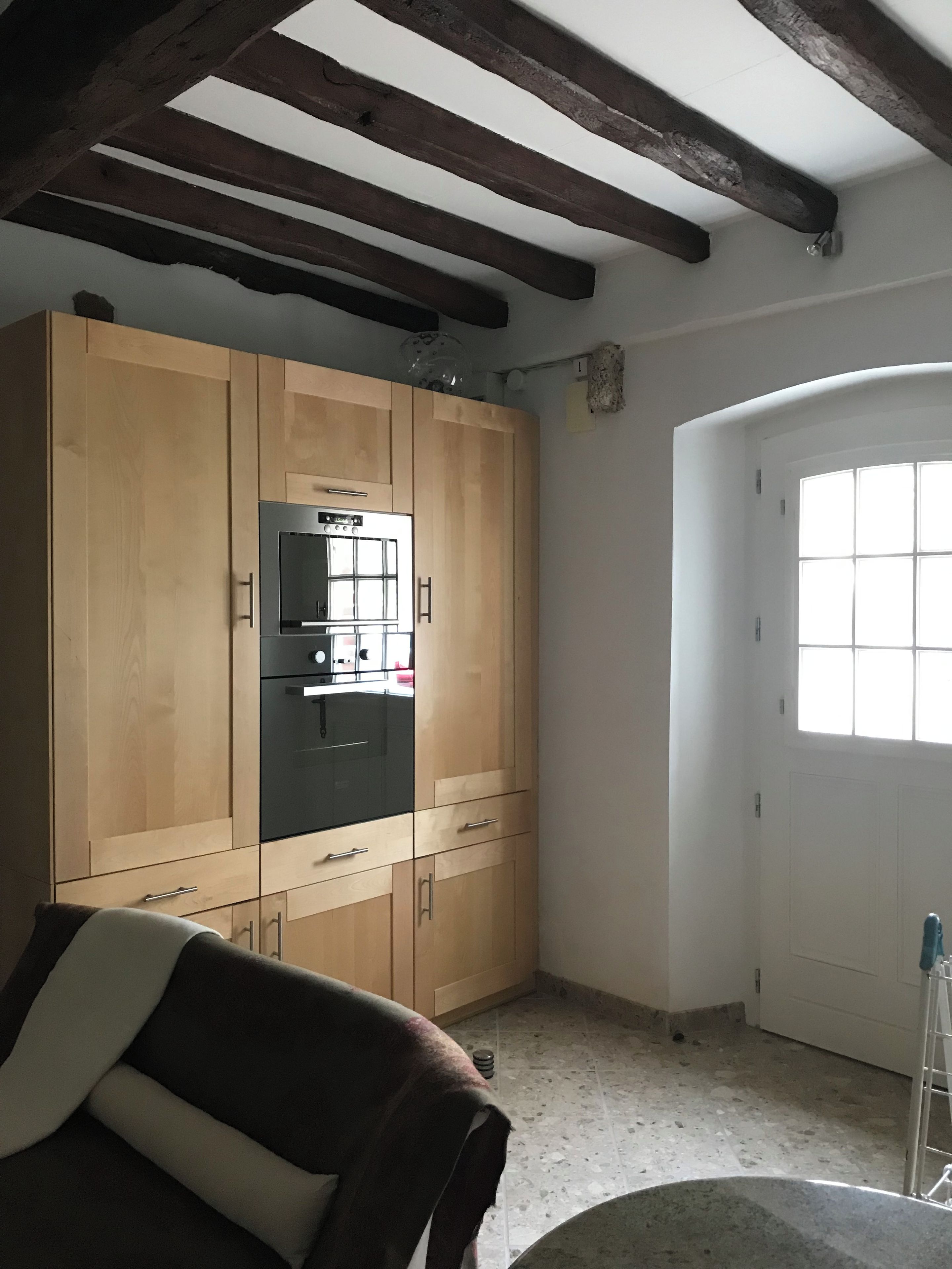 A room with a large wooden wardrobe, a built-in black refrigerator, a window with a view, and a tiled floor.