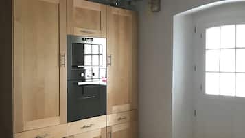 A room with a large wooden wardrobe, a built-in black refrigerator, a window with a view, and a tiled floor.