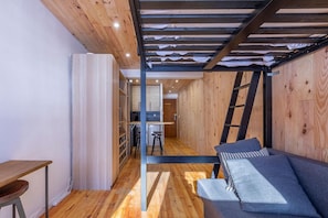 Quarto compacto com piso de madeira, um sofá com almofadas, uma pequena mesa e uma escada encostada na parede.