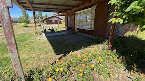 A covered outdoor area with a wooden structure, a garden with yellow flowers, and a view of a house in the background.