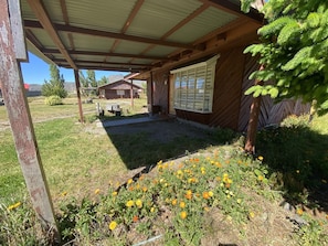 A covered outdoor area with a wooden structure, a garden with yellow flowers, and a view of a house in the background. - Patagonia Bordelago (Río Ibáñez)
