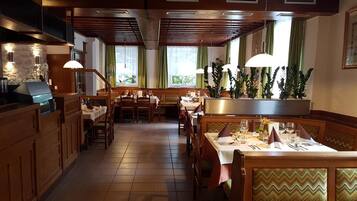 L’intérieur d’un restaurant, avec des meubles en bois, des tables dressées pour le service et de grandes fenêtres ornées de rideaux.