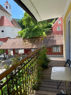 A balcony with a table and chairs, overlooking a courtyard with a building with a red roof and a white tower. - Hotel Restaurant Stv Ckl (Schwanberg)