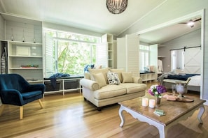 A living room with a sofa, armchair, and coffee table, with a bookshelf and a large window. - Franklin Cottage (Olinda)