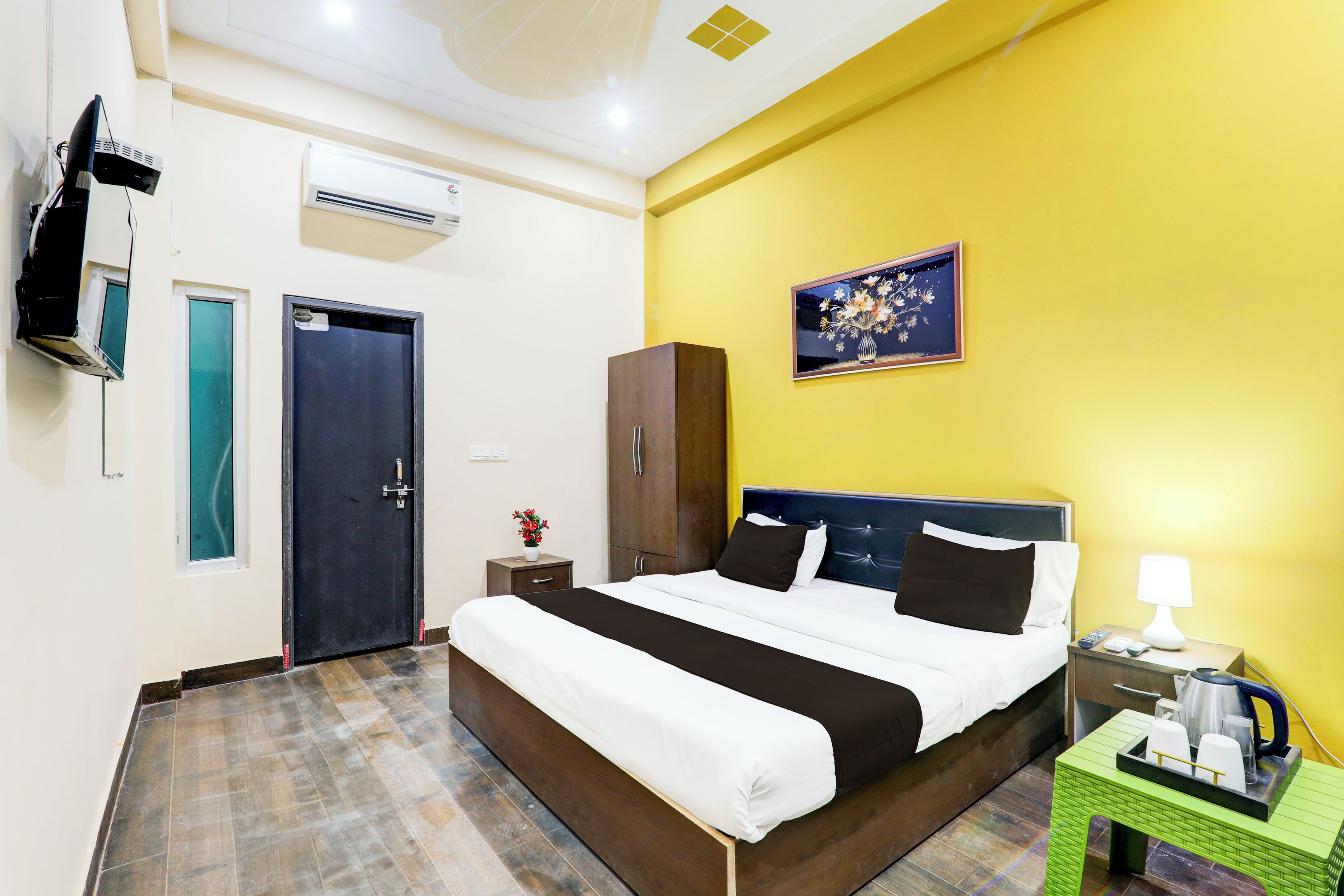 Una habitación de hotel con una cama, una mesita de noche, una pequeña mesa verde auxiliar, un cuadro en la pared y un televisor fijado a la pared.