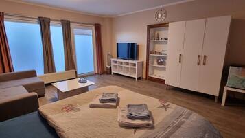 Ein Hotelzimmer mit Sofa, Bett mit Tagesdecke, Fernseher, Schrank und einem Fenster mit Vorhängen.