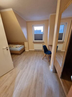 Um pequeno apartamento de um quarto com teto inclinado, uma cama, uma cadeira e duas janelas.