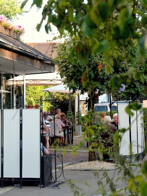 An outdoor seating area with tables and chairs, surrounded by greenery and a building with a tiled roof. - Best Breakfast - Hotel Justina (Bad Woerishofen)