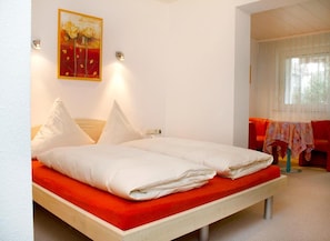 A double bed with white bedding and a red accent, a painting on the wall, and a red sofa in the background. - Best Breakfast - Hotel Justina (Bad Woerishofen)