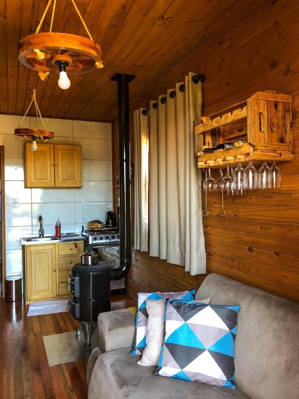 Interior acolhedor de uma cabana com teto de madeira, cozinha compacta, sofá com almofadas estampadas e espaço para vinhos.