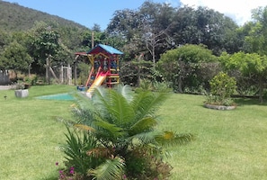 A playground with a slide and climbing structures, surrounded by lush greenery and a hill in the background. - Pousada Tia Tuquinha (Piumhi)
