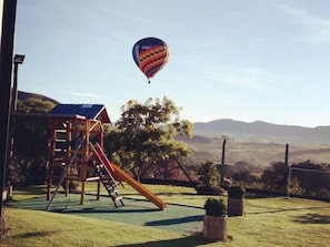 A playground with a slide and climbing structure, a hot air balloon in the sky, and a hilly landscape in the background. - Pousada Tia Tuquinha (Piumhi)