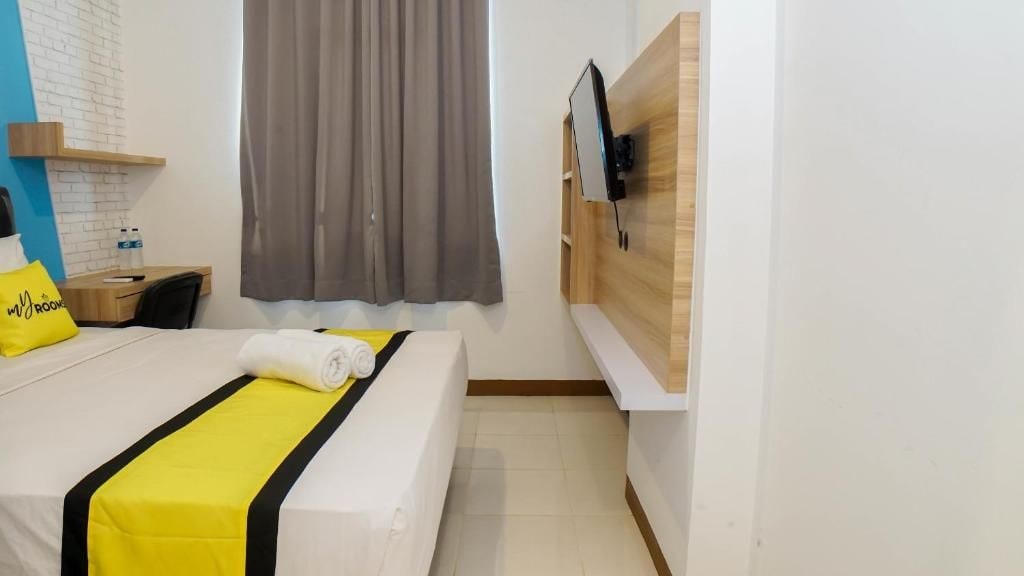 Een hotelkamer met een bed, een bureau, een stoel, een aan de muur bevestigde televisie en een raam met gordijnen.