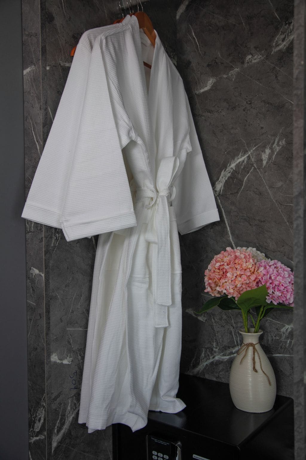 A white bathrobe hanging on a rack against a marble wall, with a vase of pink flowers on a black surface beside it.