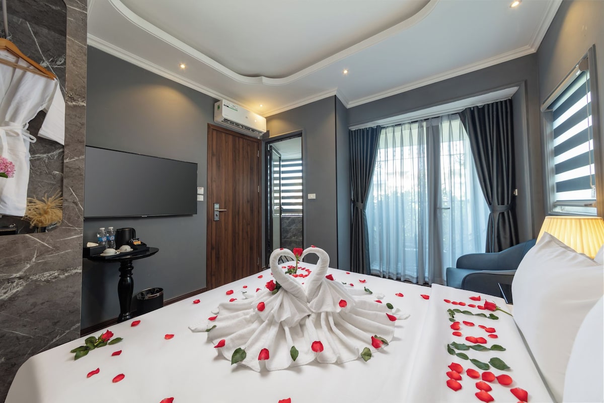 A hotel room with a bed adorned with rose petals and swan-shaped towels, a flat-screen TV mounted on the wall, a wooden door, and a window with curtains.