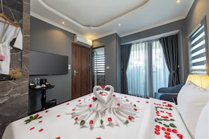A hotel room with a bed adorned with rose petals and swan-shaped towels, a flat-screen TV mounted on the wall, a wooden door, and a window with curtains. - Premium Madelis Hotel (Hanoi)