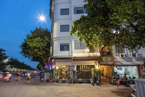 Hotel com placa indicando 'MADELIS HOTEL' e rua em frente com motocicletas estacionadas e trânsito.