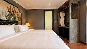 Habitación de hotel con una cama grande, una mesita de noche, una lámpara, una bata colgada en un perchero y una puerta que da a otra habitación.