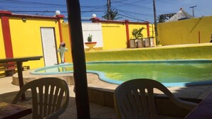 A swimming pool with a yellow wall and a door, surrounded by chairs and a table. - Top Home Pousada (Caucaia)