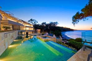 A modern pool area with a view of the ocean, surrounded by a landscaped garden and a large building with multiple levels. - Amarna Luxury Beach Resort (Nelson Bay)