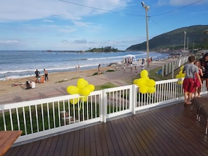 A wooden deck with a white railing overlooking a beach with people and a clear sky. - Frente Mar (Florianópolis)