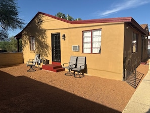 A single-story building with a red roof and beige walls, featuring two windows with white curtains, a door, and a small air conditioning unit. - Historic charming 2-bedroom house with AC in welcoming Tucson (Tucson)