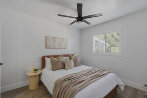A bedroom with a bed, a bedside table, a ceiling fan, a window with blinds, and a wall-mounted artwork. - LongBeach Cottage - Pool, Spa, Games, 4Miles2Beach (Long Beach)