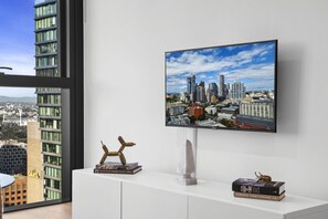 A modern interior with a large flat-screen TV mounted on a white wall, a white console table with books and a sculpture, and a view of a cityscape through a large window. - High-Rise 1-Bed Apartment in Melbourne CBD (Melbourne)