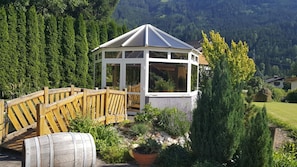 Ein hölzerner Gazebo mit Glasverkleidung, umgeben von einem Garten mit verschiedenen Pflanzen und einer Holzbrücke.
