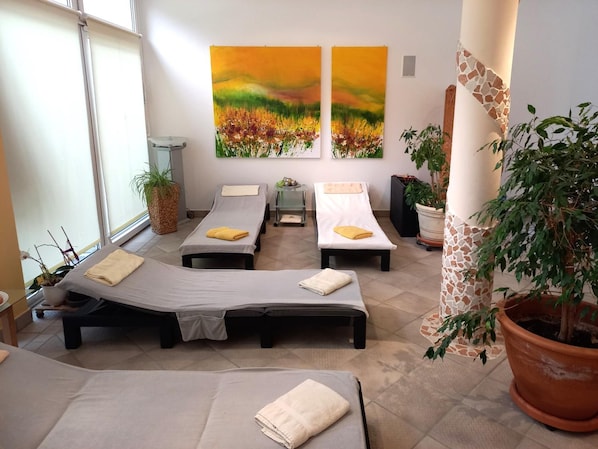 Ein Zimmer mit vier Einzelbetten, jeweils ausgestattet mit einem Handtuch und einem kleinen Tisch. An der Wand befinden sich zwei große Gemälde, ein Topfpflanze und eine Säule mit Mosaikmuster.
