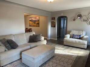 Una sala de estar con sofá, reposapiés y sillón, un cuadro en la pared y un pasillo que conduce a una puerta.