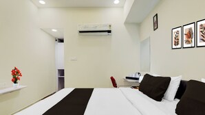Ein Hotelzimmer mit Bett, Schreibtisch, Stuhl, einem roten Sessel, einer weißen Vase mit Blumen, einer Klimaanlage und gerahmten Kunstwerken an der Wand.